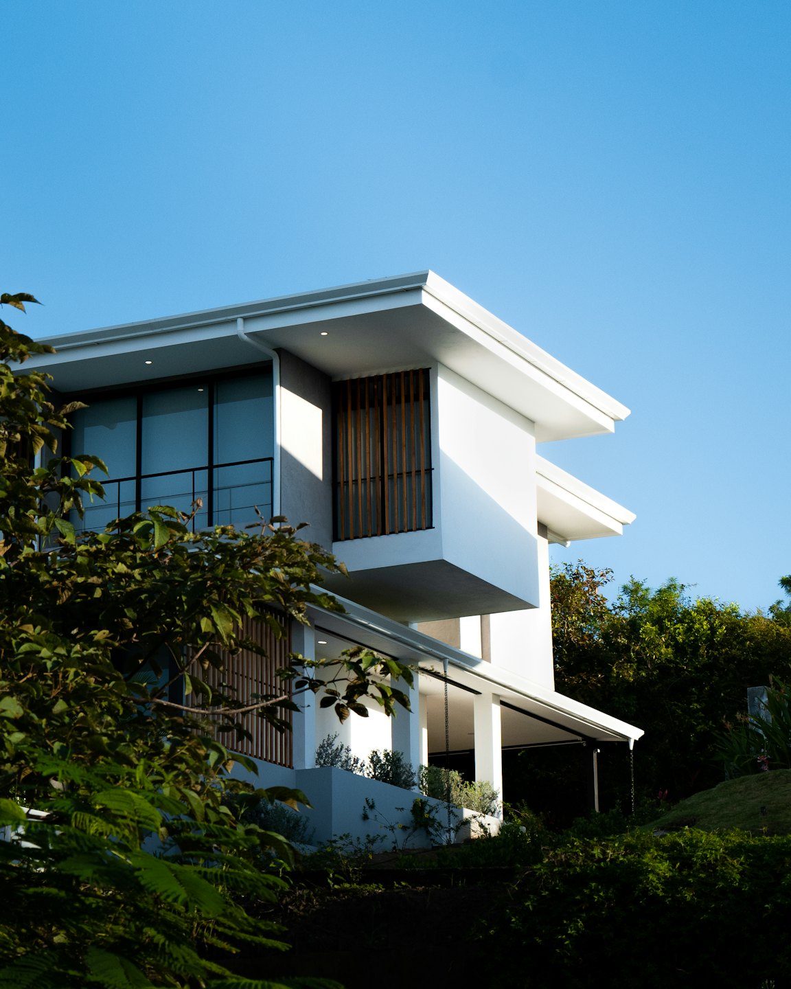 Modern white house with wooden accents behind green trees.